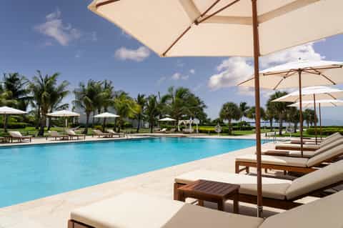Resort pool area with lounge chairs, umbrellas, palm trees, and manicured grounds