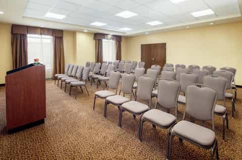 Meeting room with rows of gray padded chairs facing a wooden podium with beige walls and brown curtains