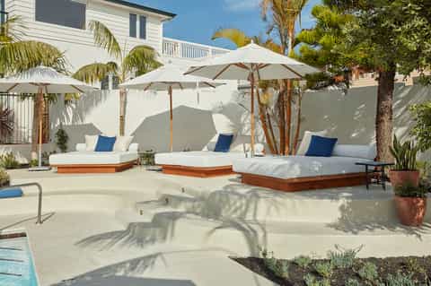 Residential pool deck with lounge beds, umbrellas, tropical plantings, and modern white home