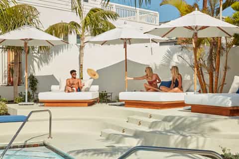Beach club lounge area with white cushioned daybeds under cream umbrellas, palm trees, and clear sky