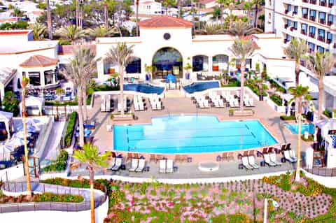 Aerial view of resort with rectangular swimming pool, white lounge chairs, palm trees, and Spanish-style central building
