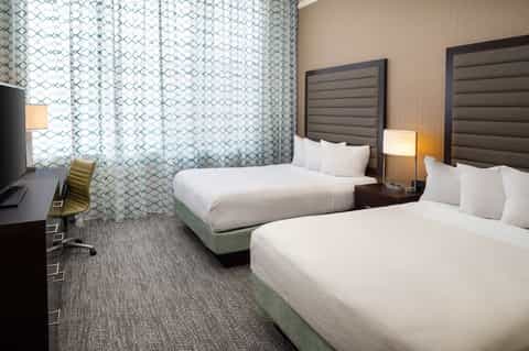 Double queen hotel room with patterned curtains, bedside lamps, and work desk