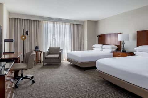 Hotel guest room with two beds, work desk, armchair, and balcony access
