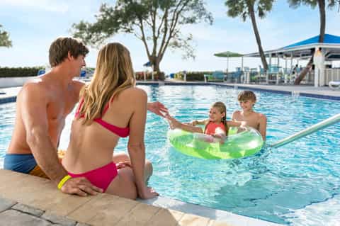 Family enjoying resort pool with children playing in inflatable ring, parents supervising from deck