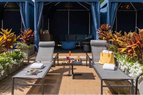 Upscale rooftop lounge with lounge chairs, blue cabanas, flowering plants, and city views