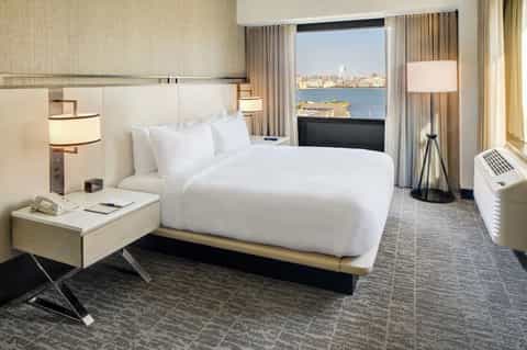 Modern hotel bedroom with queen bed, waterfront city views, floor-to-ceiling windows, and contemporary furnishings