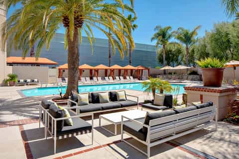 Resort pool area with palm trees, lounge seating, orange umbrellas, and modern architecture