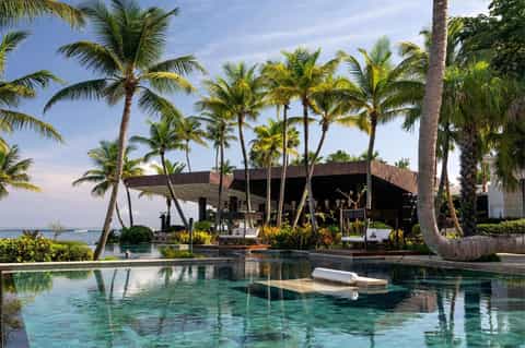 Luxury oceanfront pool with palm trees, modern dark building, and crystal-clear turquoise water at tropical resort