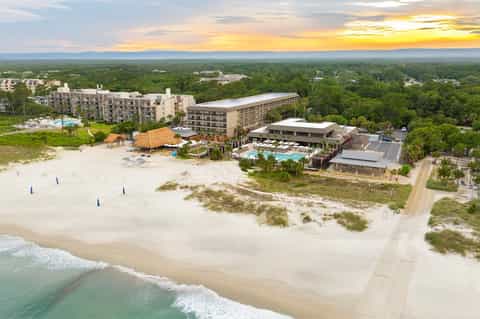 Aerial view of beachfront resort with pools, buildings, sandy beach, and sunset over coastal landscape