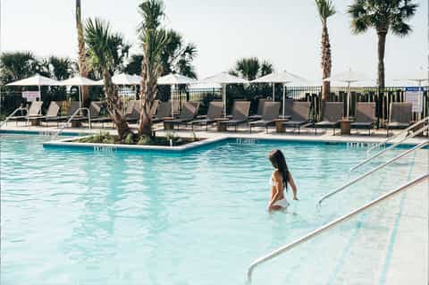 Tropical resort pool with palm trees, white umbrellas, lounge chairs, and guest swimming