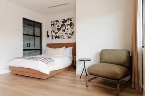 Modern hotel bedroom with wooden bed frame, earth-tone chair, and minimalist decor