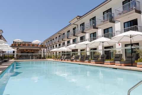 Outdoor pool with light blue water, white umbrellas, lounge chairs, and multi-story hotel building
