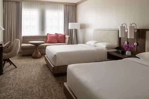 Modern hotel room with two queen beds, upholstered headboard, sitting area with sofa, and floor-to-ceiling curtains