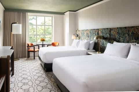 Bright hotel bedroom with two queen beds, patterned headboard, large windows, and modern furnishings