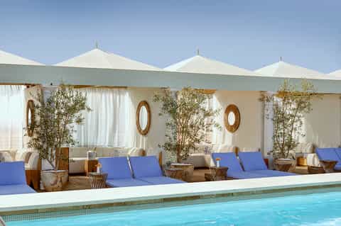 Luxury poolside lounge with cream umbrellas, blue loungers, olive trees, and elegant pergola structure