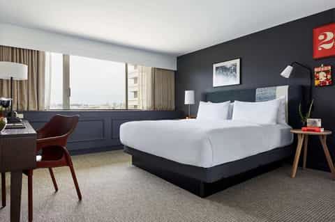 Contemporary hotel bedroom with dark accent wall, white bedding, floor-to-ceiling windows, and city skyline view