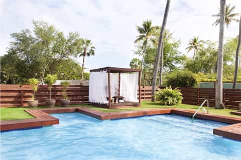 Residential pool with wood decking, brown cabana with white curtains, palm trees, and fenced tropical setting