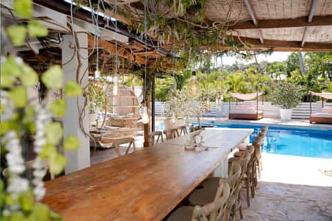 Covered poolside dining area with wooden table, hanging plants, and blue pool view