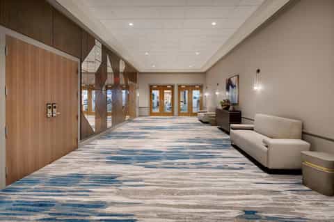 Modern hotel hallway with wooden doors, neutral walls, blue patterned carpet, and seating areas