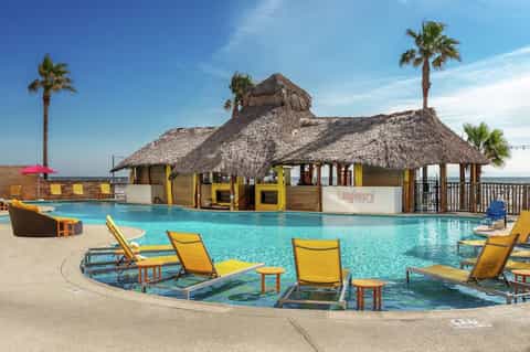Resort pool with thatched-roof bar, palm trees, yellow lounge chairs, and clear blue water