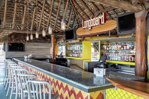 Beachfront tiki bar with 'Longboard' sign, thatch roof, colorful decorations, and full liquor shelves
