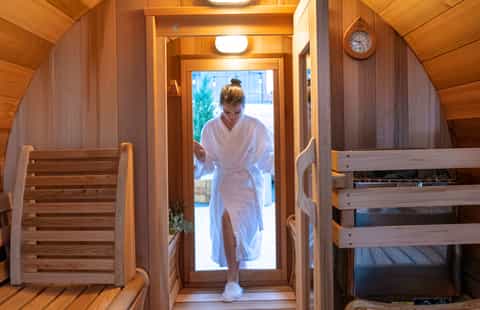 Wooden sauna interior with woman in white robe entering through glass door