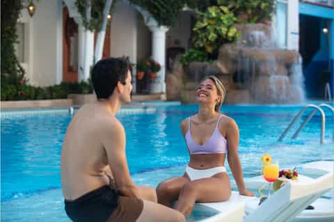 Couple enjoying pool at hotel with waterfall feature, tropical architecture, and blue umbrellas
