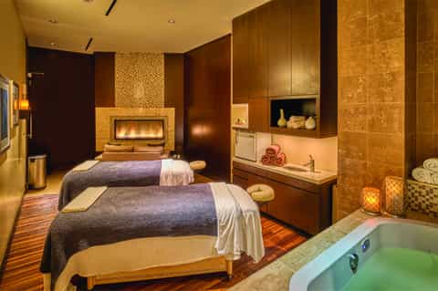 Luxury spa treatment room with fireplace, two massage beds, and bronze tile accents