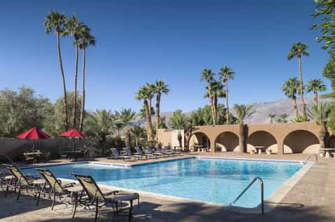 Desert resort pool with red umbrellas, lounge chairs, arched structures, and palm trees
