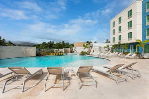 Resort pool area with turquoise water, tan lounge chairs, hotel building, and mountain backdrop