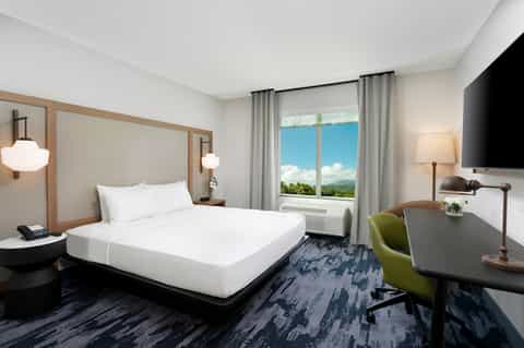 Modern hotel bedroom with queen bed, desk area, and large window overlooking mountains and blue sky