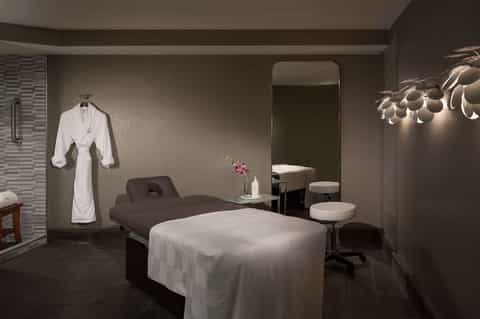 Modern spa treatment room with massage bed, robe, white towels, mirror, and contemporary lighting fixtures