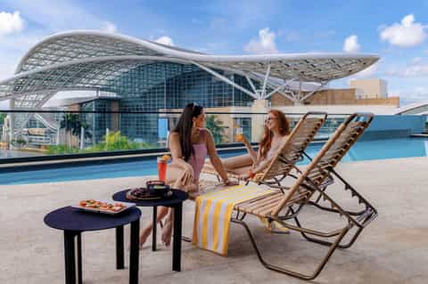 Two women relaxing on lounge chairs by a pool with modern architecture and water features in background