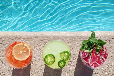 Three colorful tropical cocktails with citrus and herbs on poolside concrete deck