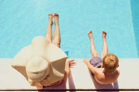 Two people dangling legs over bright blue pool edge wearing sun hats