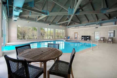 Indoor heated pool facility with bright blue water, wooden table, exposed ceiling beams, and large windows