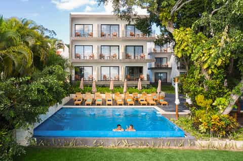 Resort courtyard with swimming pool, lounge chairs, umbrellas, and multi-story building surrounded by tropical greenery