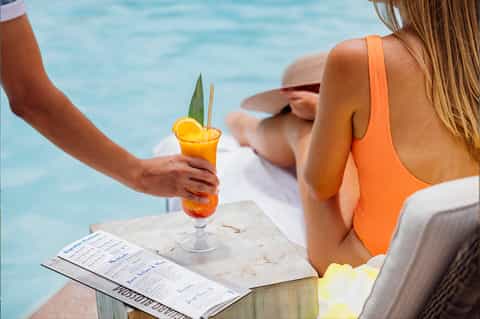 Woman in orange bikini holding tropical cocktail poolside with menu and ocean view in background