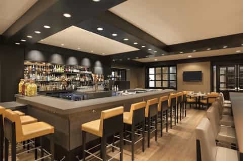 Modern bar lounge with dark wood, yellow leather stools, bottles, recessed ceiling lights, and seating area
