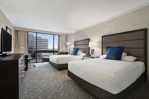 Modern hotel room with two queen beds, taupe headboards, white bedding, blue accent pillows, and city skyline views through floor-to-ceiling windows