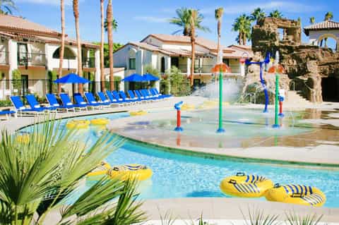 Resort pool with splash pad, blue loungers, and Spanish-style buildings surrounded by palm trees