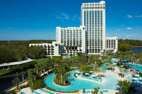 Aerial view of large white resort with curved towers, multiple pools, and palm trees surrounded by greenery