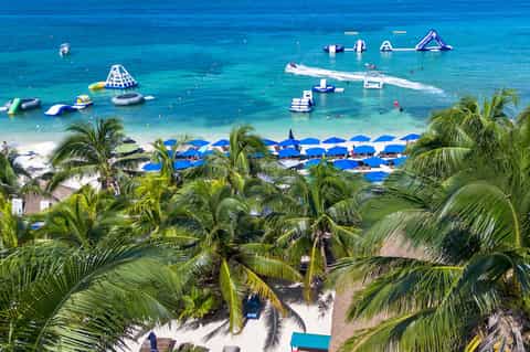 Aerial view of tropical beach with blue umbrellas, water sports equipment, palm trees, and clear turquoise ocean