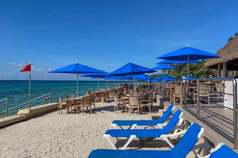 Beach club with blue umbrellas, lounge chairs, tables, and ocean view with red flag