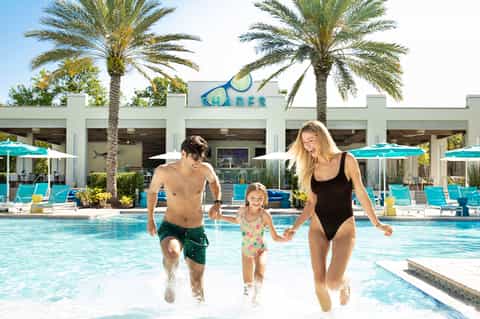 Family running into resort pool with palm trees and turquoise umbrellas under clear sky