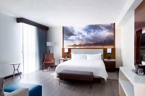 Contemporary hotel bedroom with king bed, dramatic cloud artwork, wooden headboard, floor-to-ceiling windows, and modern furnishings