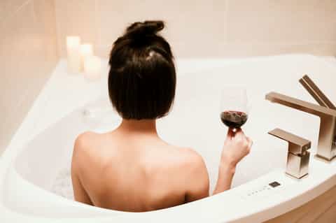 Woman relaxing in white bathtub holding red wine with modern chrome faucet and candles