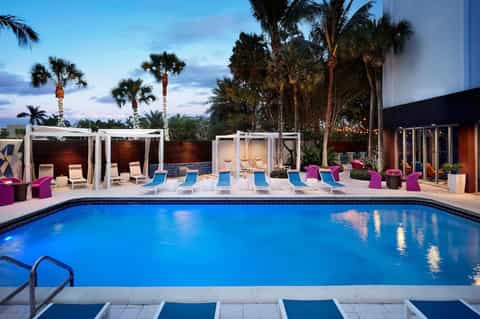Resort pool at twilight with palm trees, modern cabanas, colorful seating, and building facade