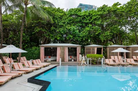 Modern resort pool with striped loungers, white umbrellas, cabanas, and lush green backdrop