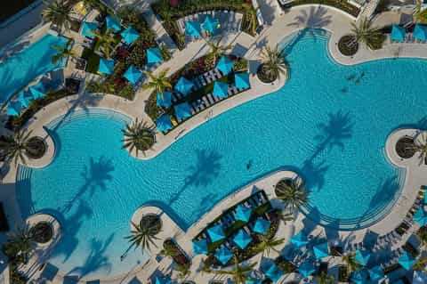 Aerial view of expansive resort pool complex with turquoise water, blue umbrellas, palm trees, and lounge chairs on surrounding deck